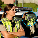 Police officer in a high-visibility vest with a camera on a car, surrounded by police vehicles.